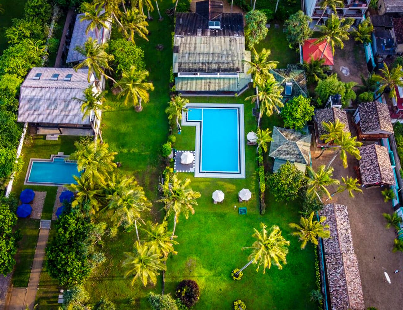 Vista aérea de un resort con una piscina central, rodeado de palmeras y varios edificios.