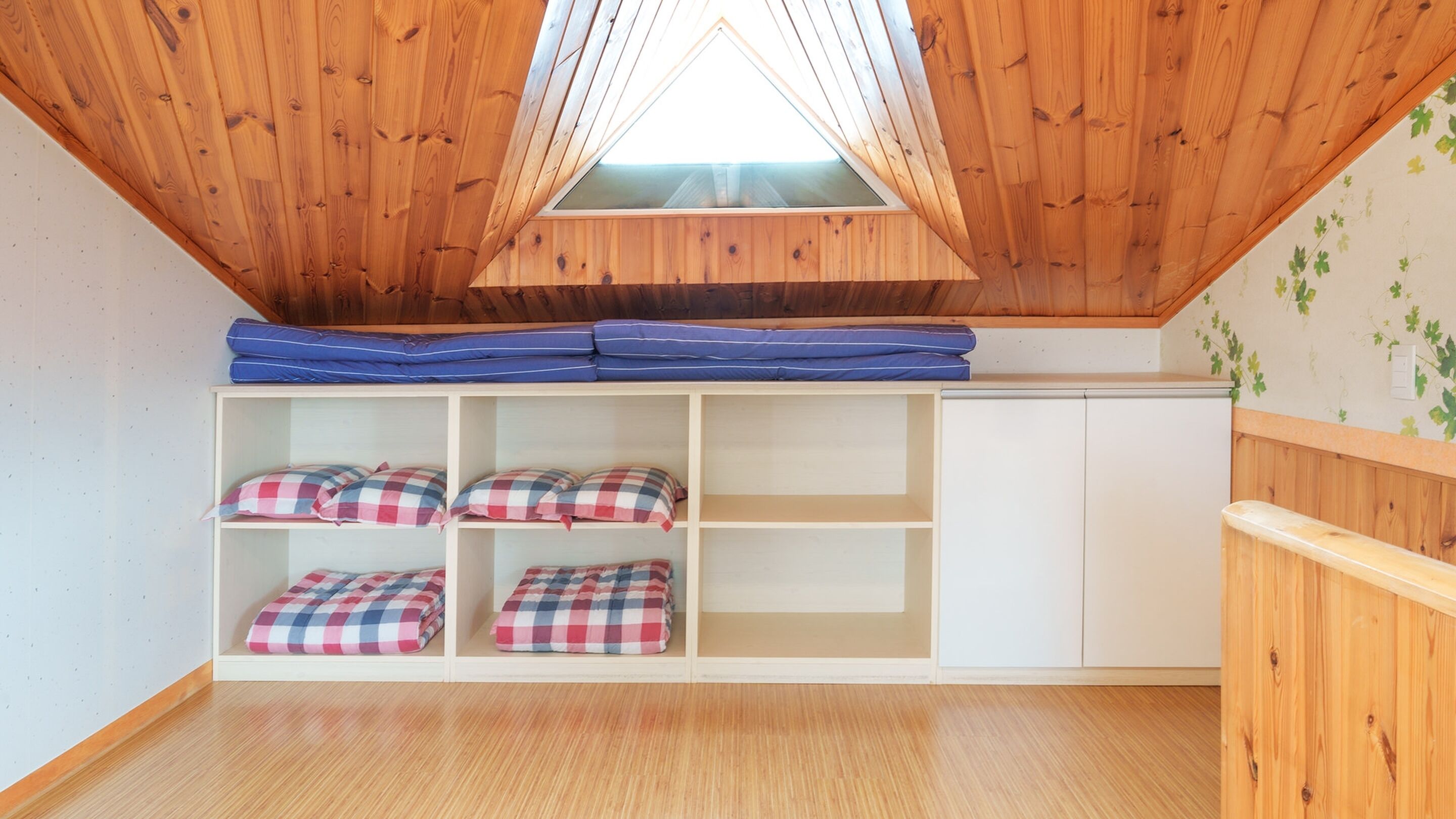 A room with a sloped ceiling, wooden walls, and a skylight. There are bunk beds with blue mattresses and checkered pillows.