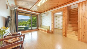 A spacious room with a wooden floor, a bench, a table with a vase of flowers, and a large window offering a view of greenery and hills. - Gangwha Jeongwon Pension (Incheon)