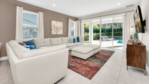 A modern living room with a sectional sofa, a wooden cabinet, and a large window offering a view of a pool and greenery.