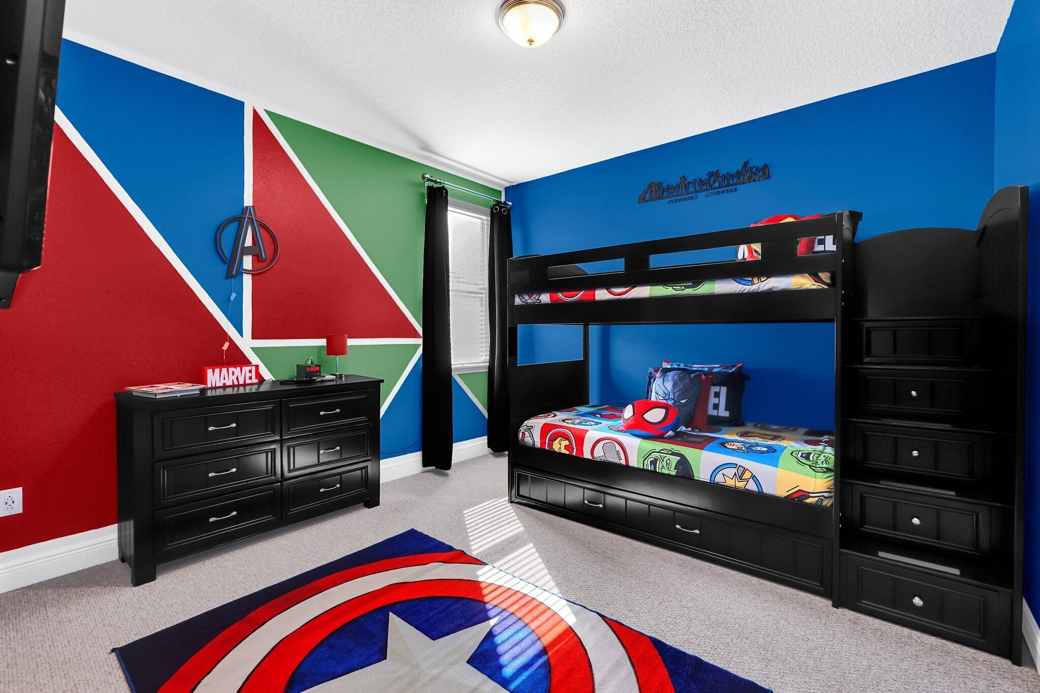 Een kamer met een stapelbed, een zwarte dressoir, een blauwrode muur en een kleed met een Marvel thema.