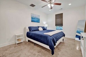 A bedroom with a bed, a ceiling fan, a nightstand, a lamp, and a window with a view of a car.