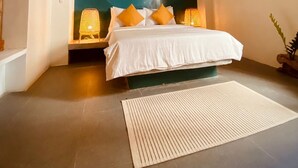 A bedroom with a bed, a ceiling fan, a wall with a sunset painting, and a nightstand with a lamp.