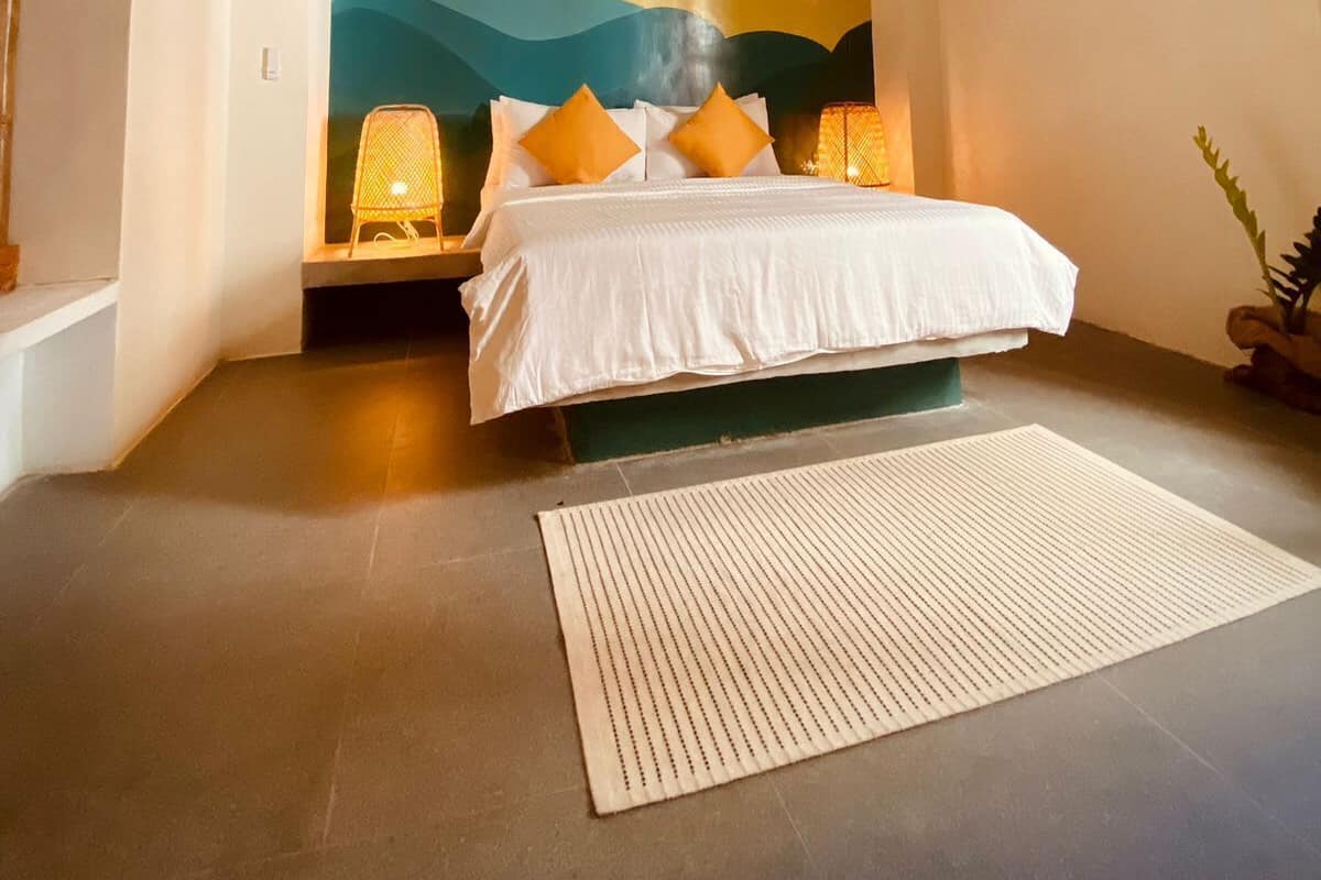 A bedroom with a bed, a ceiling fan, a wall with a sunset painting, and a nightstand with a lamp.