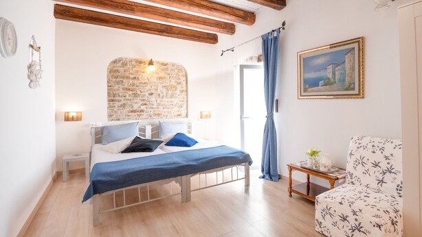 A bedroom with a stone wall, wooden beams, a bed with blue bedding, a chair, and a painting of a coastal scene. - Penelope Stone House (Corfu)