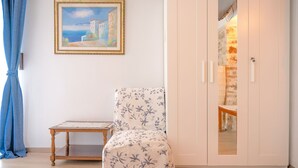 A room with a floral-patterned chair, a small wooden table, a framed painting, and a white wardrobe with a mirror. - Penelope Stone House (Corfu)