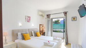 A hotel room with two beds, a bedside table, a lamp, a TV, and a window with curtains. - Angela Apartment in Kassiopi (Corfu)