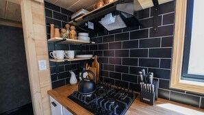 A compact kitchen with black tiled backsplash, wooden shelves, and a gas stove. - Chipmunk Tiny Home at The Woolly Bugger Inn (Estes Park)