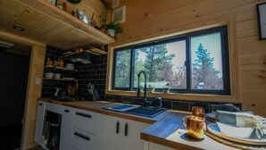 A kitchen with a large window, wooden cabinets, and a countertop with a kettle and a mug. - Chipmunk Tiny Home at The Woolly Bugger Inn (Estes Park)