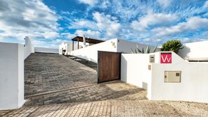 Un sendero pavimentado que conduce a una entrada con puerta de madera, paredes blancas y un cielo azul despejado.
