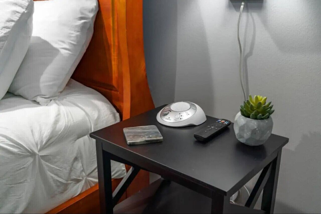 A neatly made bed with white bedding and orange curtains, a bedside table with a white lamp, a remote control, and a book.