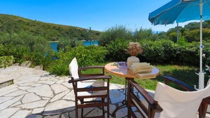 A stone patio with a table and chairs, a blue umbrella, and a view of a lake and hills. - Spiros Saint Stephanos (Corfu)
