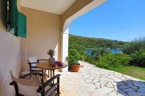 A terrace with a table and chairs, a potted plant, and a view of a hillside and water body. - Spiros Saint Stephanos (Corfu)