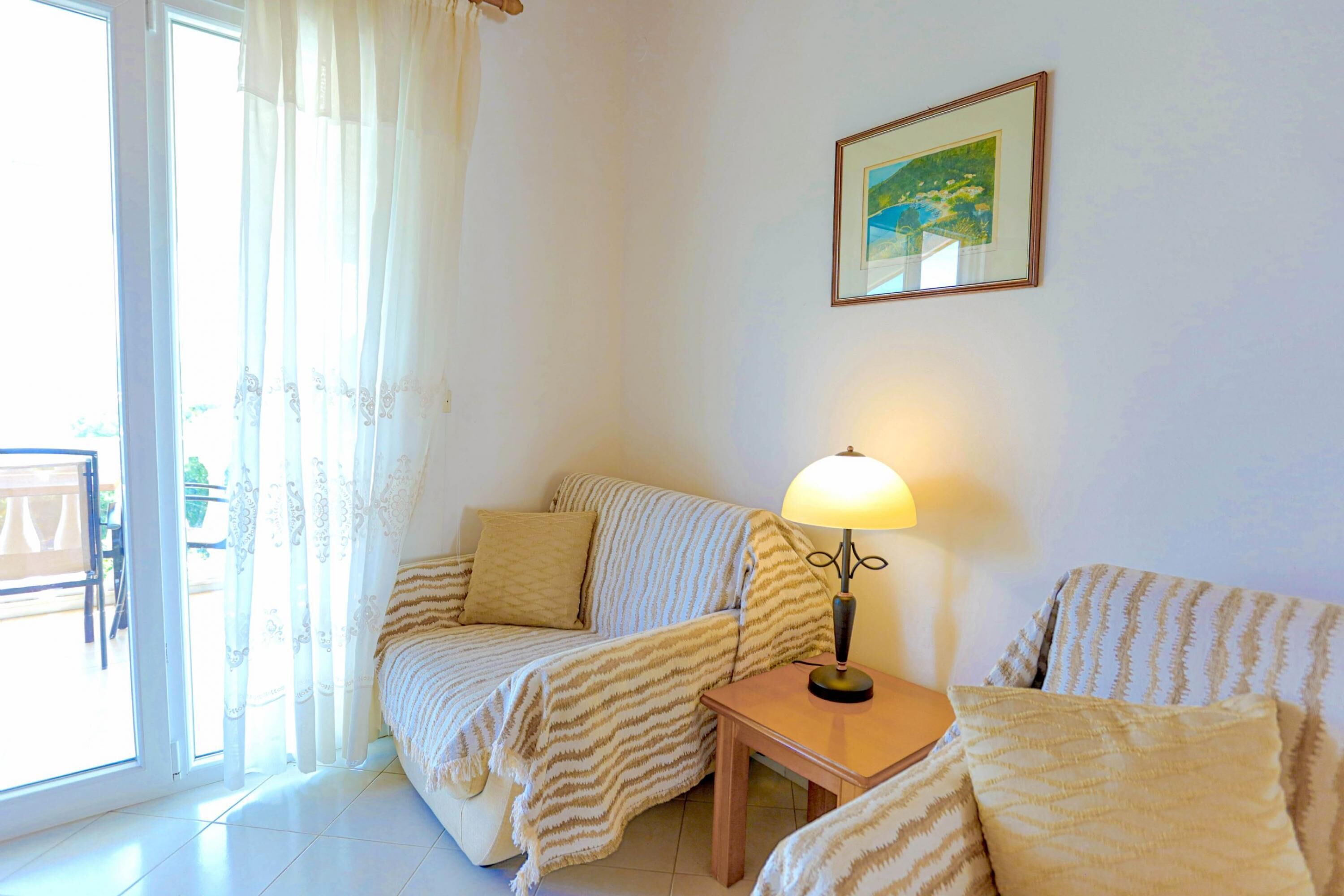 A room with a sofa, a small table with a lamp, a framed picture on the wall, and a view through a window.