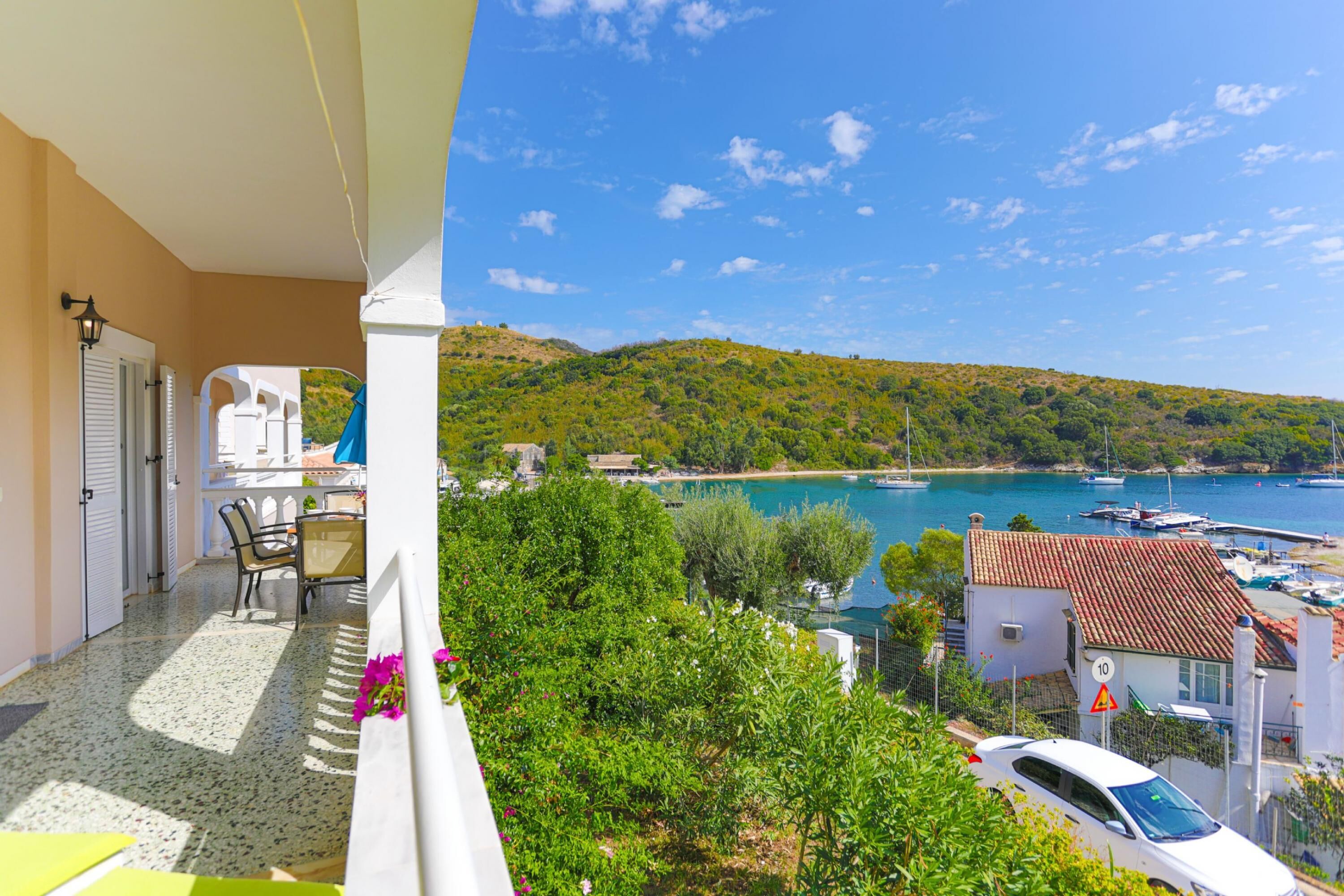 A balcony with a view of a coastal town, a dining area with chairs, and a white car parked nearby.