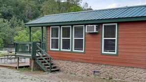 A rustic cabin with a stone foundation, a green porch with a staircase, and a metal roof. - Cabin 11 - Beautiful cabin with a queen, full and twin bed. Great for days outdoors and nights in. (Connellsville)