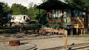 A rustic cabin with a green roof and wooden deck, surrounded by gravel and trees. - Cabin 11 - Beautiful cabin with a queen, full and twin bed. Great for days outdoors and nights in. (Connellsville)