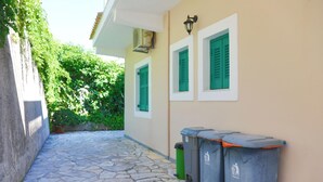 A narrow stone-paved pathway leading to a building with green shutters and trash bins. - Anna Apartment Agios Stephanos (Corfu)