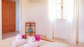 A hotel room with a wooden wardrobe, a small wooden shelf with books, a window with white curtains, and two rolled towels with pink flowers. - Anna Apartment Agios Stephanos (Corfu)