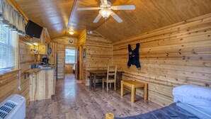 Una cabaña acogedora con interior de madera, un ventilador de techo, una cocina con pileta y estufa, una mesa de comedor con sillas, y una cama con colchón.