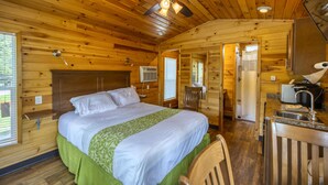 A wooden cabin bedroom with a bed, a ceiling fan, a desk with a sink, and a bathroom visible through an open door. - Cabin 3 - Everything you need in this Cabin for a great weekend. (Connellsville)