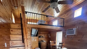 Eine Holzhütte mit einem Schlafzimmer auf dem Dachboden, ausgestattet mit einem Bett, einem Schreibtisch und einem Stuhl. Eine Treppe führt zum Dachboden, und an der Wand befindet sich eine Klimaanlage.