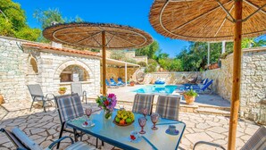 An outdoor patio with a pool, stone walls, and a table set with chairs and umbrellas. - Villa Divista in Paxi (Paxos)