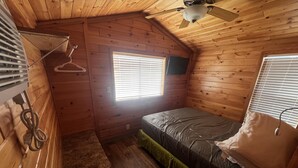 Een kleine houten kamer met een enkel bed, een plafondventilator en twee ramen met zonwering.