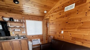 Interior acollidor d'una cabana amb un sòl de fusta, un sofà, una cuina amb un vàter i armaris, i un ventilador de sostre.