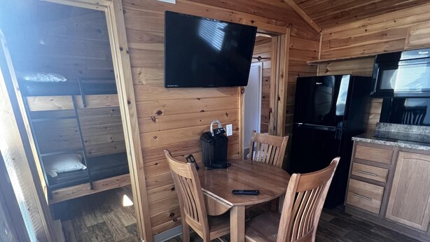 Interior d'una cabana de fusta amb taula de dinar, cadires, televisió de pantalla plana, nevera negra i microones.