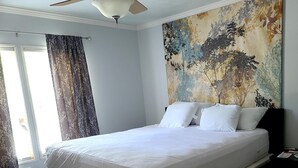Ein Schlafzimmer mit einem Bett, einem Deckenventilator, einem Fenster mit Vorhängen und einer Wand mit einem Baum-Wandbild.