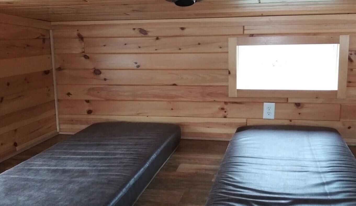 A small cabin with two beds, wooden walls, a window, and a power outlet.