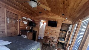 A compact wooden cabin with a bed, a small kitchenette, a dining area, and a TV mounted on the wall.