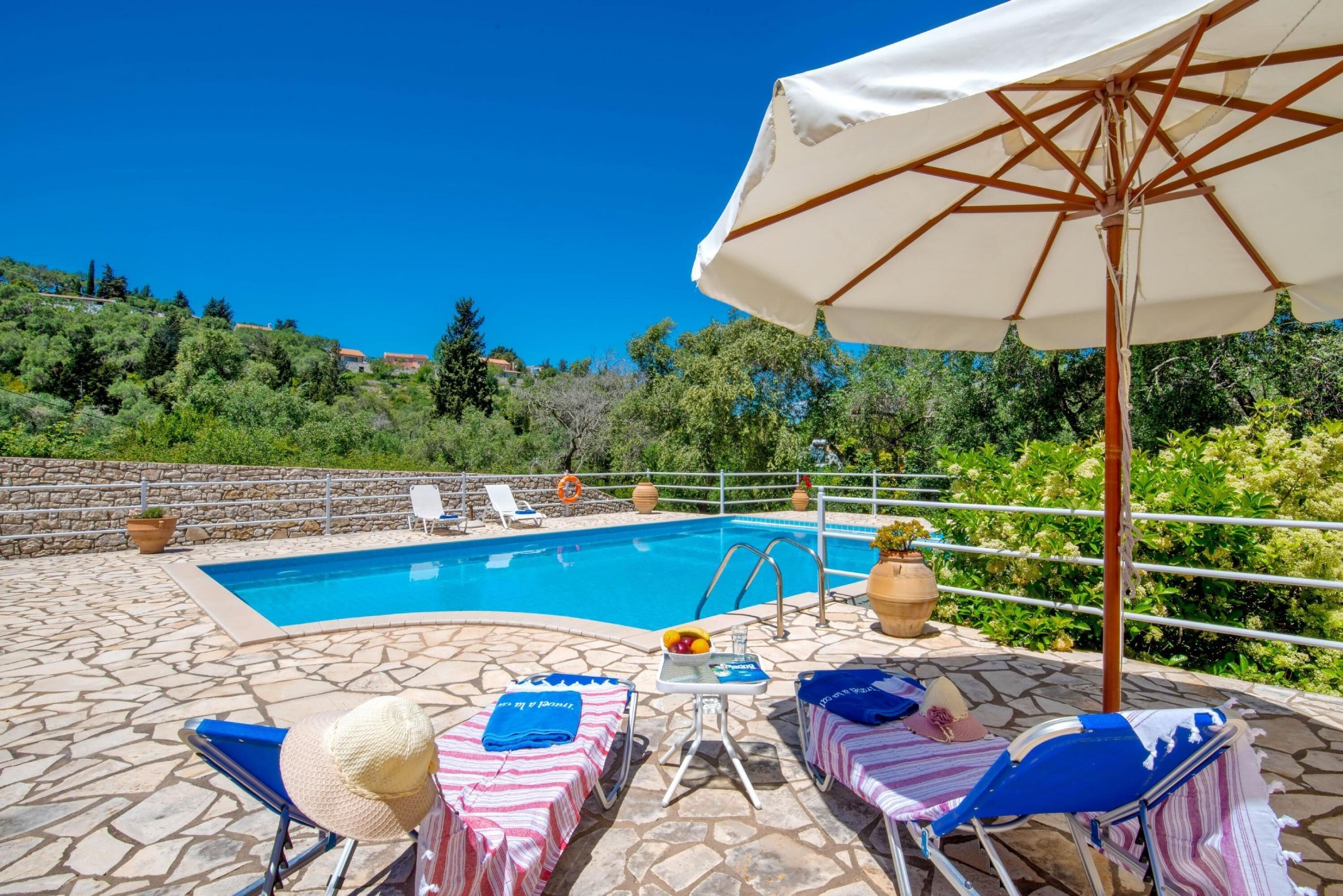 Zona de piscina amb gandules, una taula amb un plat de fruita i un parasol gran.
