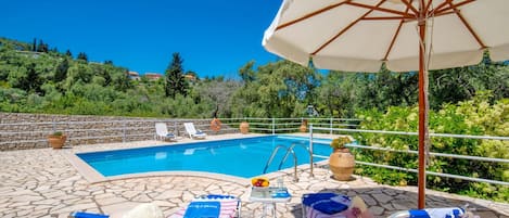 Zona de piscina amb gandules, una taula amb un plat de fruita i un parasol gran.