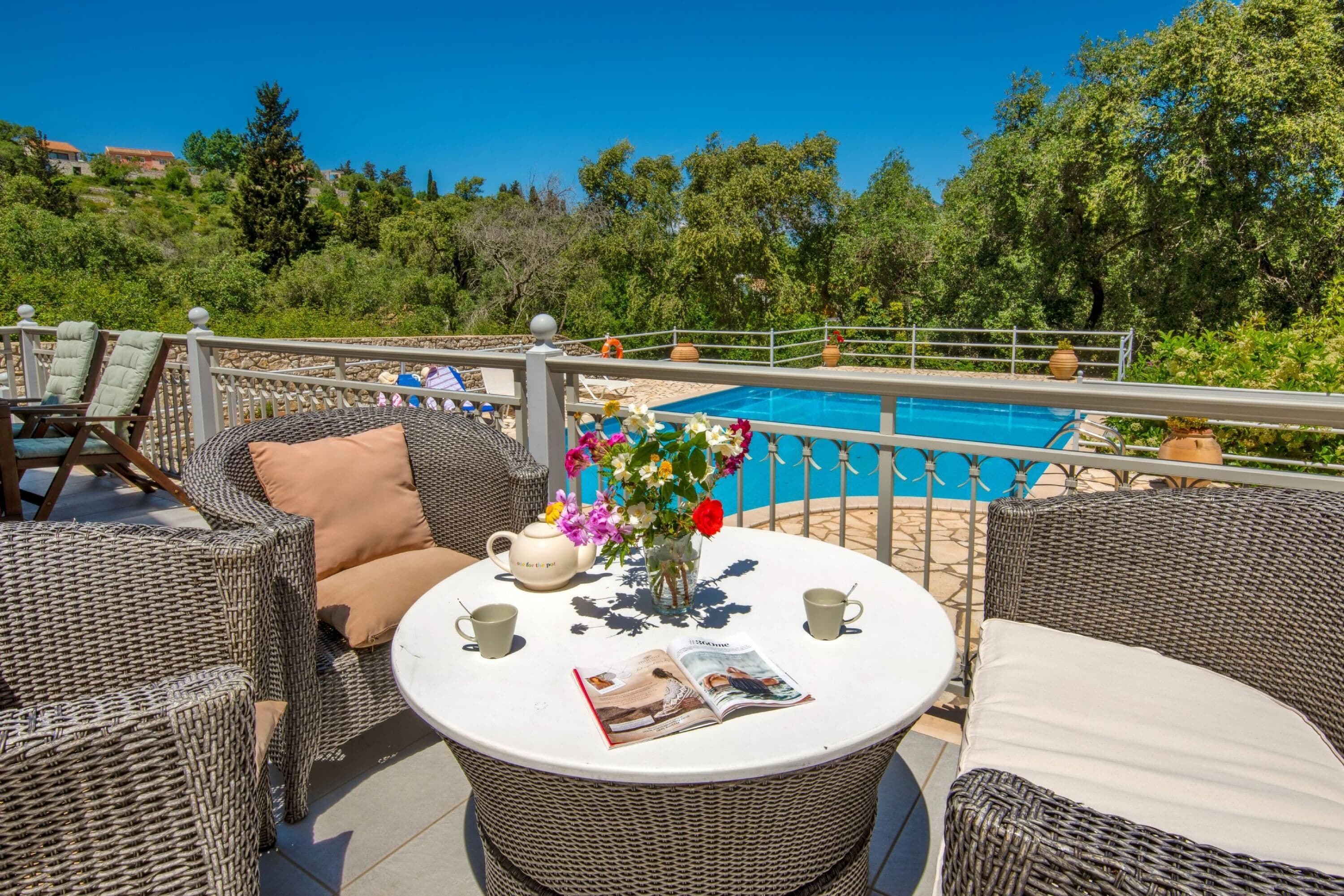 A wicker outdoor seating area with a table, chairs, a vase with flowers, a teapot, and a magazine. A pool is visible in the background.