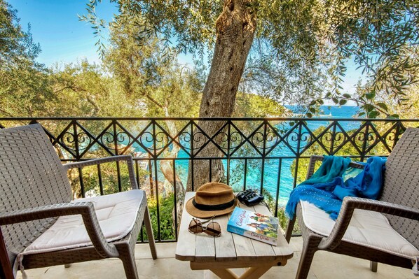 A balcony with wicker chairs, a table with a hat, sunglasses, and a book, overlooking a turquoise sea and green foliage.
