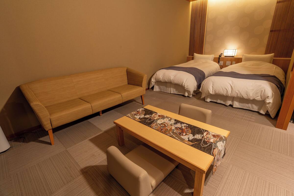 A hotel room with a sofa, a bed with two pillows, a bedside table with a lamp, and a patterned table runner.