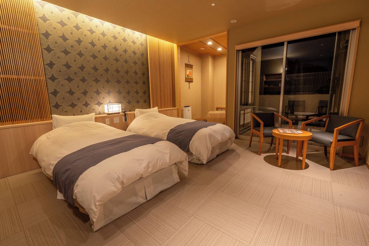 A hotel room with two beds, a small table with chairs, a nightstand, and a view of the outside balcony.