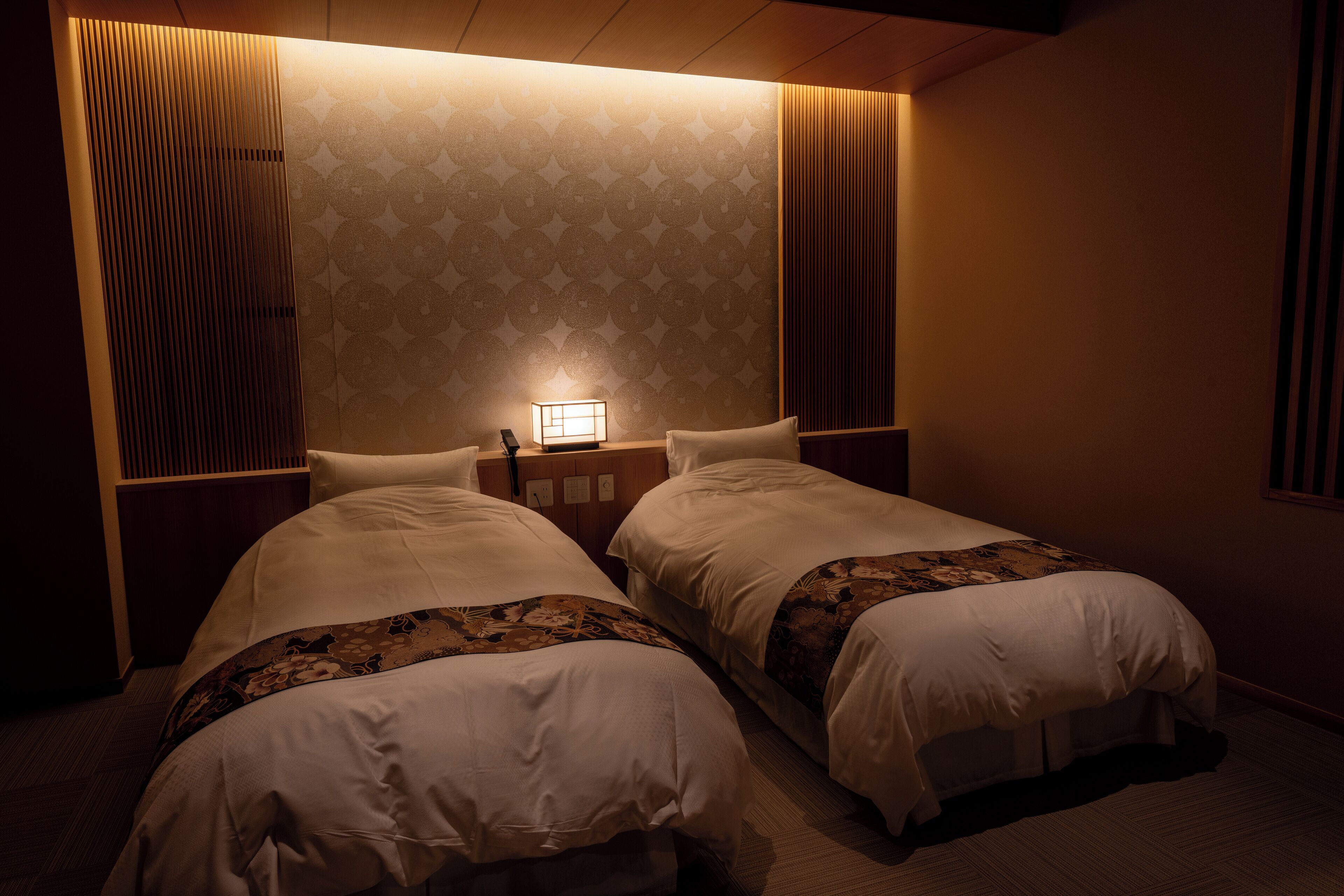 Habitación de hotel con dos camas, una mesita de noche con lámpara y un papel tapiz estampado.