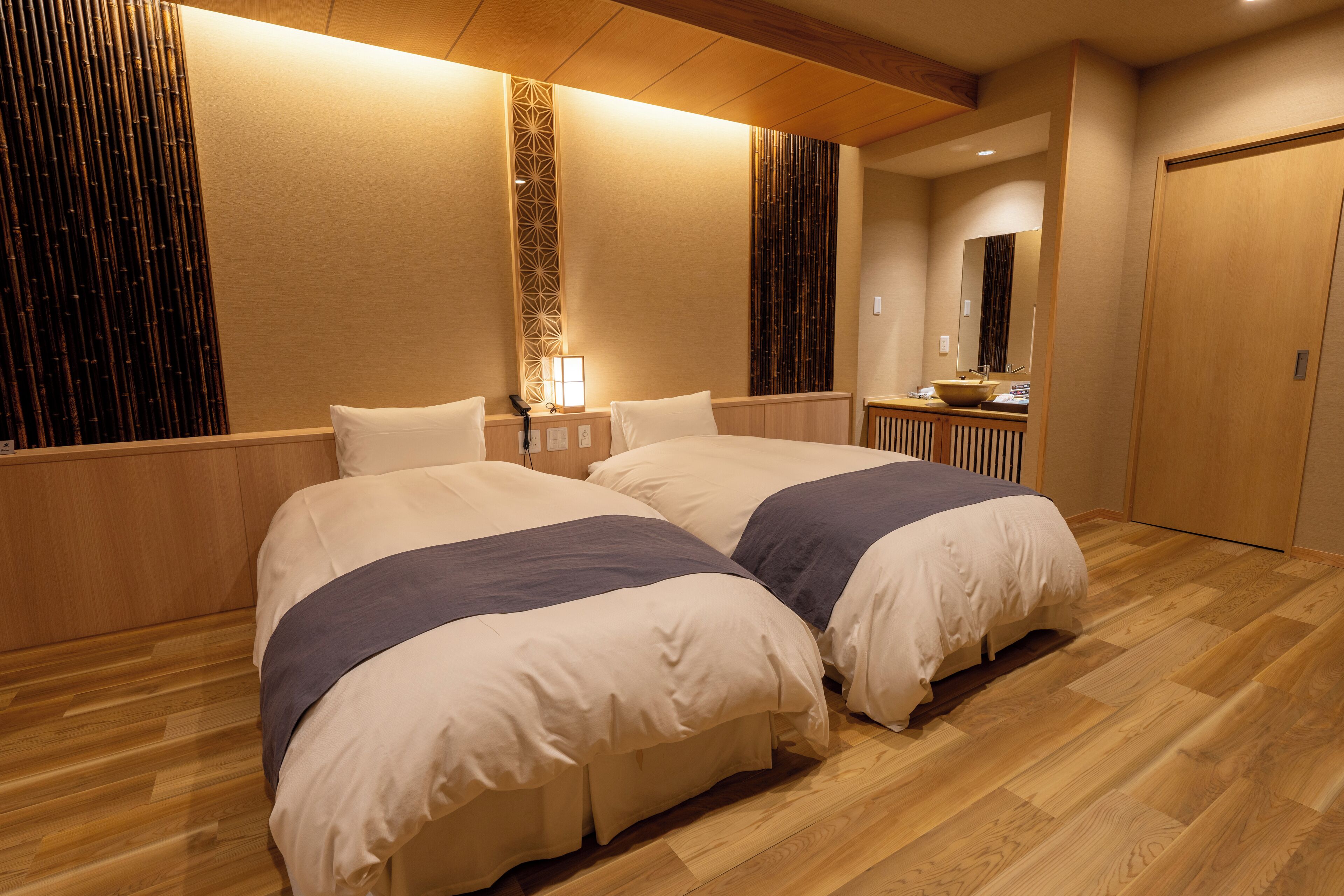 Quarto de hotel com duas camas, piso de madeira, mesa de cabeceira com abajur e um espelho grande.