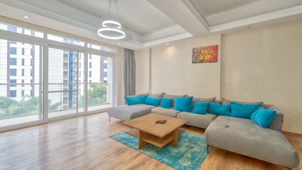 A modern living room with a grey sectional sofa, a wooden coffee table, and a large window offering a city view. - Kikambala Modern Apartment with Balcony (Nairobi)