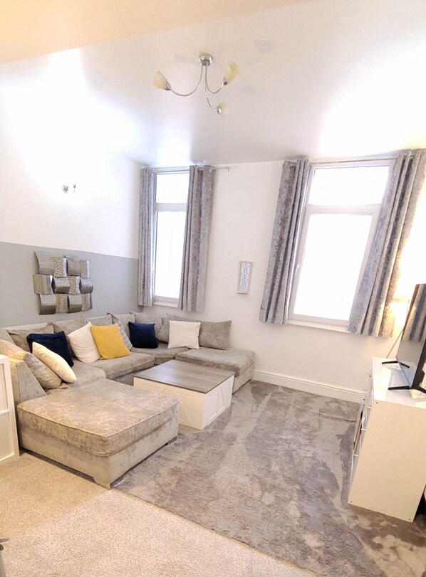 A living room with a sectional sofa, a coffee table, and a desk with a computer. There are two windows with curtains, a chandelier, and a wall-mounted artwork.