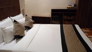 A neatly made bed with white linens and a black and beige striped bed runner. There is a wooden desk with a chair and a small shelf with bottles and a kettle. - I Towne (Deoghar)