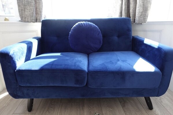 A blue velvet sofa with a round cushion in a room with wooden flooring and a window with curtains. - West Lake District Stays -contractor Accommodation (Maryport)