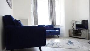 Ett vardagsrum med en blå soffa, en tv-apparat på ett skåp och ett fönster med gardiner.