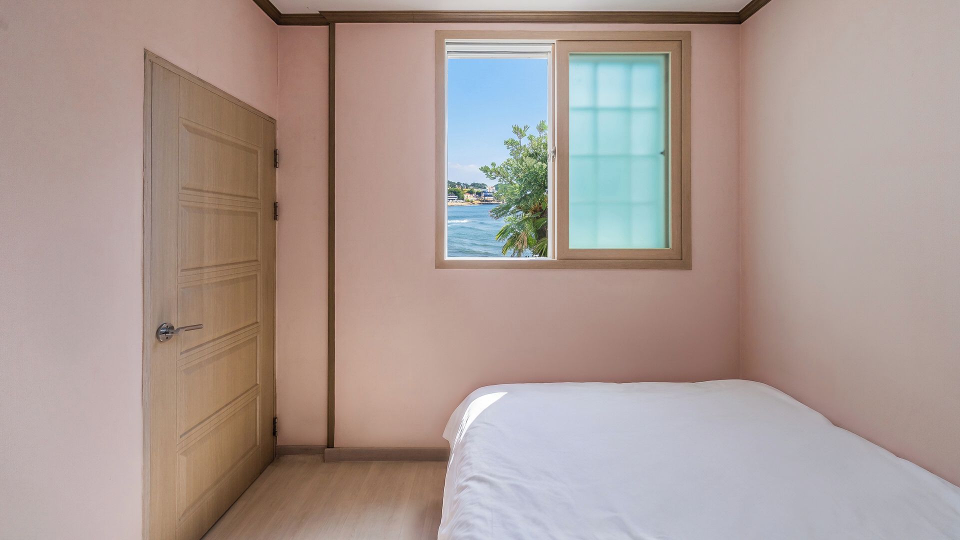 Une chambre avec un lit simple, une fenêtre donnant sur la mer et une porte en bois.