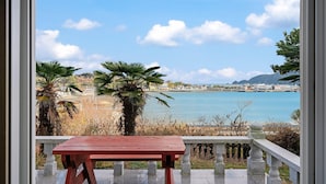 Un balcon avec une table rouge, donnant sur une zone côtière où l’on peut admirer une eau d’un bleu limpide et des bâtiments situés au loin.