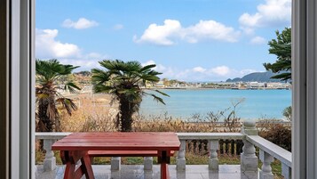 Un balcón con una mesa roja, con vista a una zona costera de aguas azules cristalinas y edificios a la distancia.