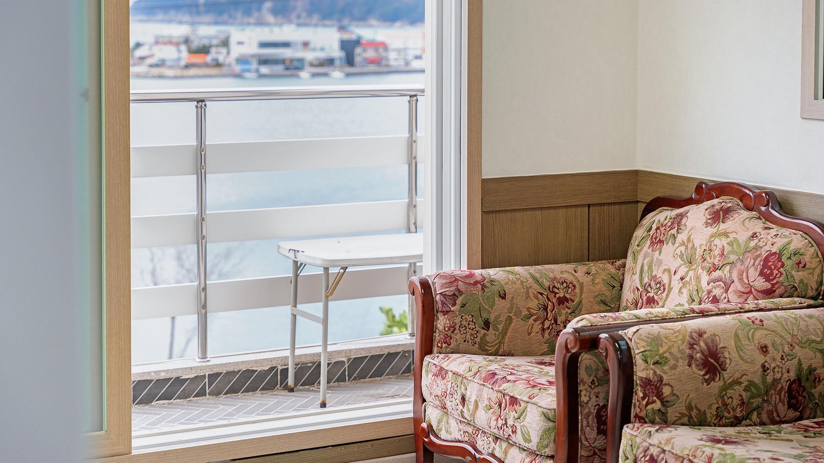 Una habitación con vistas a un puerto deportivo, un sillón de madera con tapicería floral y una mesita junto a la ventana.